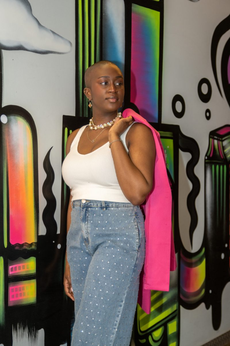 Junia Janvier in a white top and jeans, holding a pink blazer over her shoulder by a colorful mural