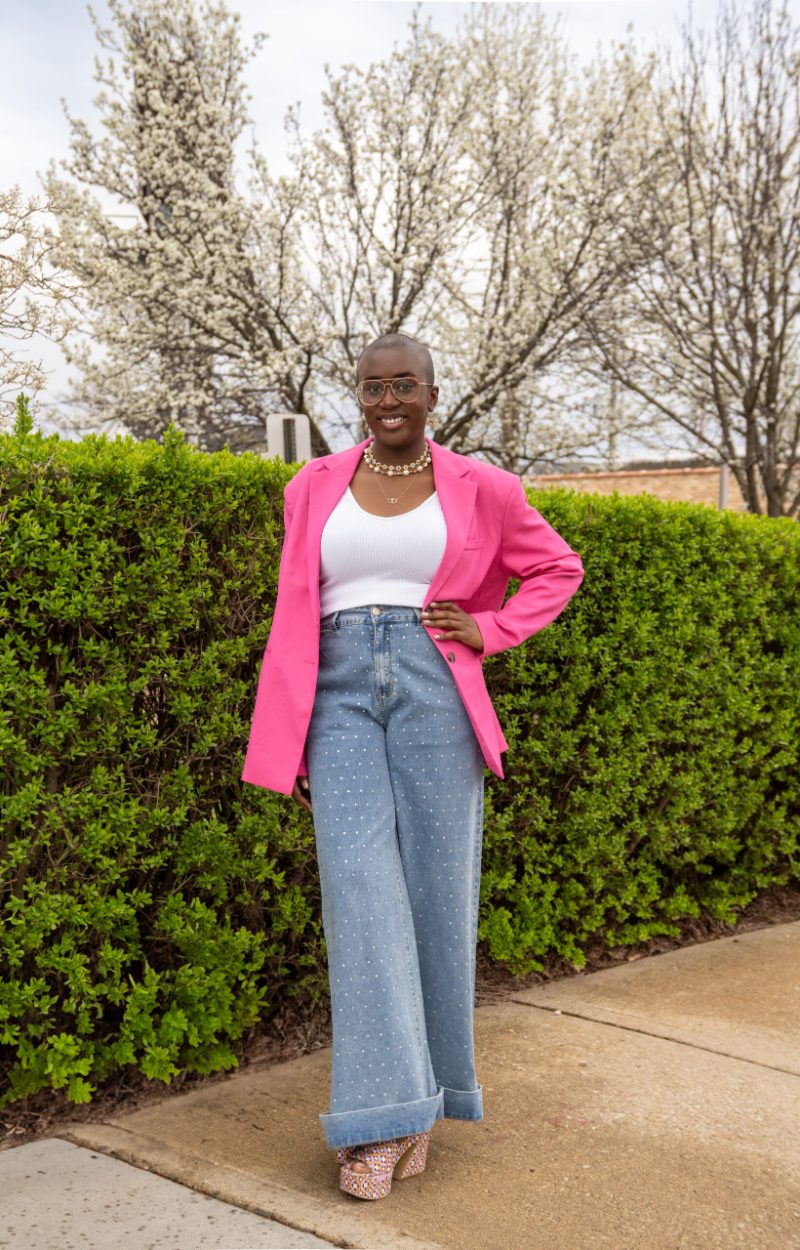 Junia Janvier smiling in a pink blazer and jeans in front of a green hedge