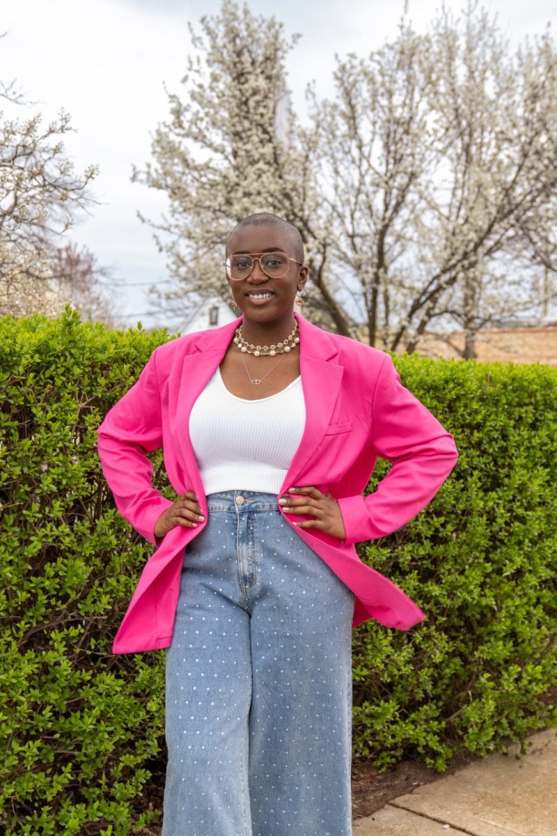 Junia Janvier in a pink blazer and white top, hands on hips, in front of a green hedge